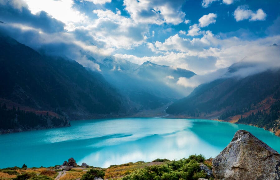 Turquoise alpine lake surrounded by snow-capped mountains and forested valleys under blue sky with white clouds
